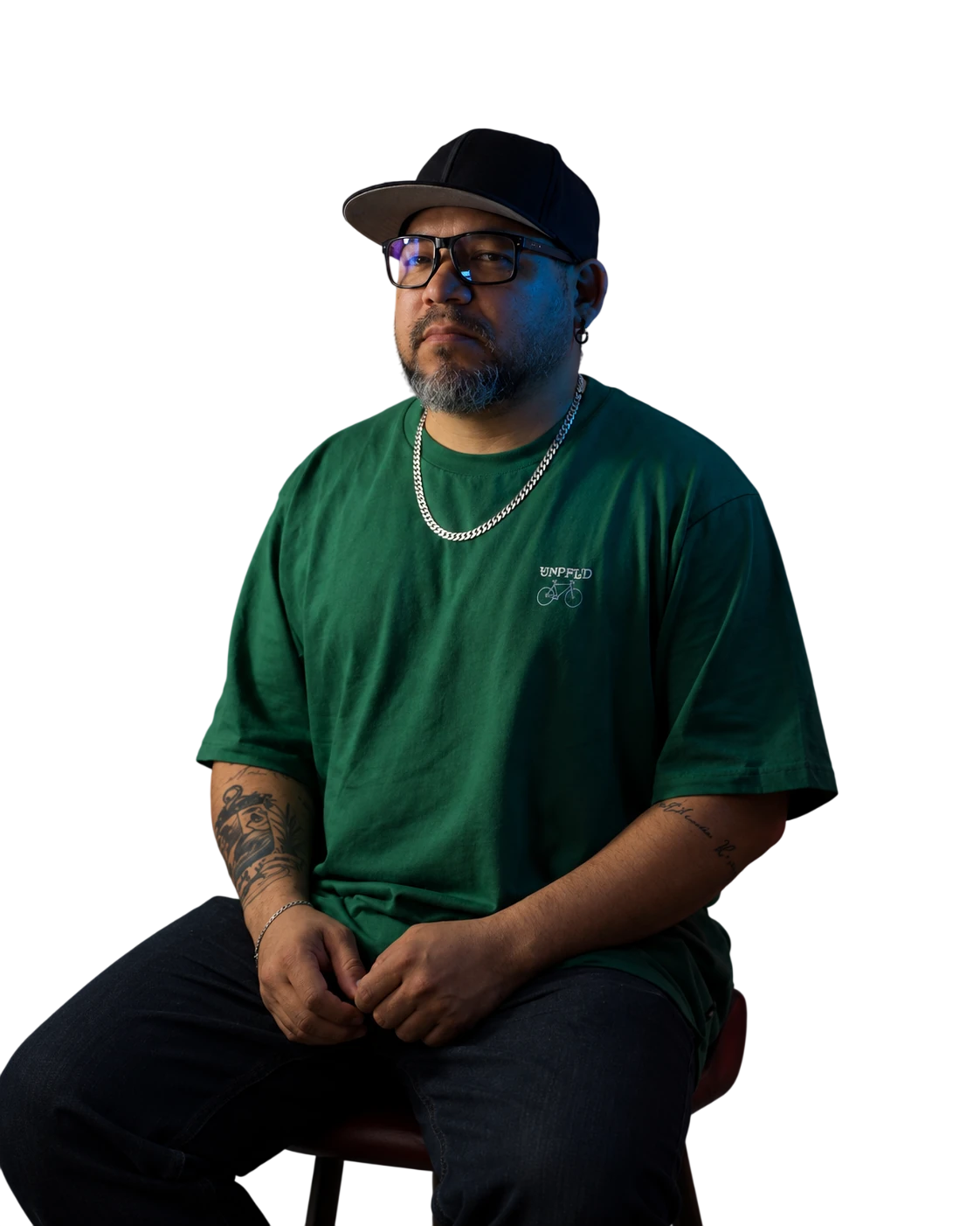 Retrato de Freidel Velásquez con gorra negra, gafas y camiseta verde, sentado en un taburete
